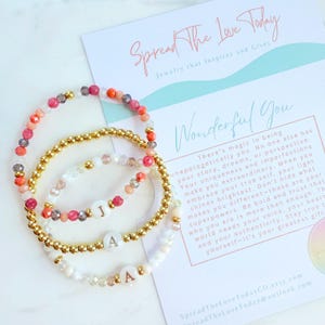 Puede incluir: Tres pulseras de cuentas con detalles dorados y abalorios con iniciales. Las pulseras presentan una mezcla de cuentas de colores en tonos rosa, naranja y blanco. Una tarjeta con el texto "Spread The Love Today" y "Wonderful You" está en el fondo.