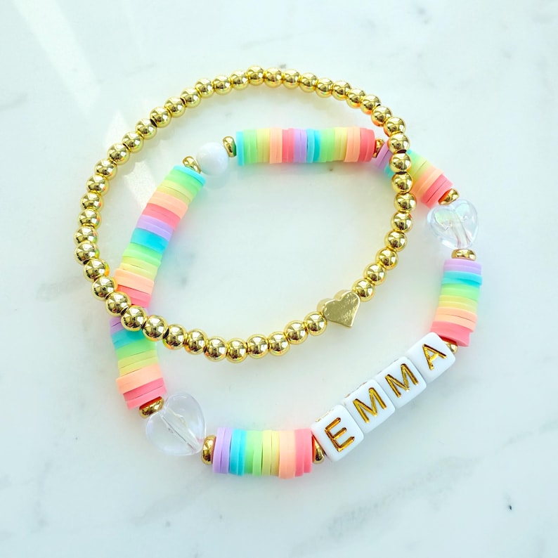 May include: Two bracelets, one with small gold beads and the other with rainbow-coloured flat beads. The name "EMMA" is spelled out in white letter beads. Gold heart charms and clear heart-shaped beads are also featured in the design.