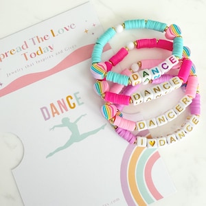 May include: Set of four colorful bracelets with the words "DANCE", "DANCER", "I LOVE DANCE" and "DAN" spelled out in white letters. The bracelets are made of pink, purple, blue and white beads with rainbow heart charms.