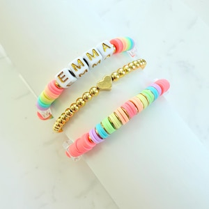 May include: Three colourful bracelets. One bracelet features the name "EMMA" in gold letters on white blocks, with rainbow-coloured disc beads. Another is gold beaded with a heart charm. The third bracelet has pastel disc beads.
