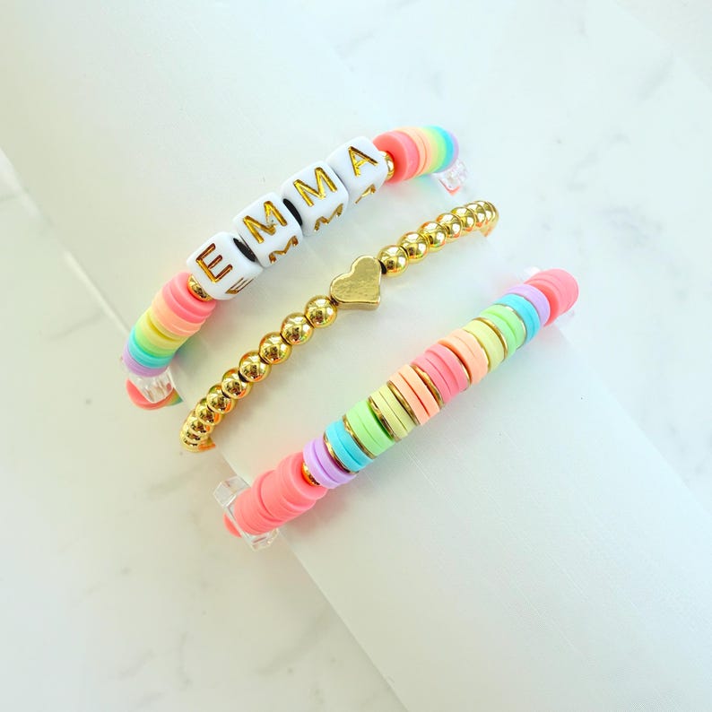 May include: Three colourful bracelets. One bracelet features the name "EMMA" in gold letters on white blocks, with rainbow-coloured disc beads. Another is gold beaded with a heart charm. The third bracelet has pastel disc beads.