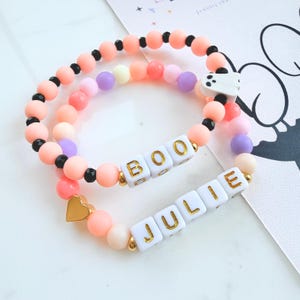 Op de afbeelding: Twee elastische armbanden met matte en glanzende kralen in perzik, roze, paars en zwart. Eén armband bevat witte kubuskralen met de tekst BOO en een spookbedel, de andere heeft witte kubuskralen met de tekst JULIE en een goudkleurig hartje.