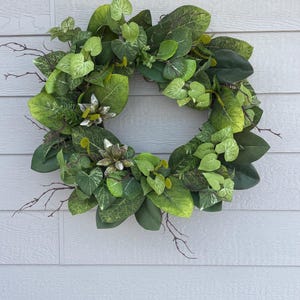 May include: A vibrant green wreath composed of various leaf shapes and textures, including large, glossy leaves and smaller, patterned ones. Twig-like branches are interspersed throughout, and small, white floral accents add a touch of contrast. The wreath is a decorative item.