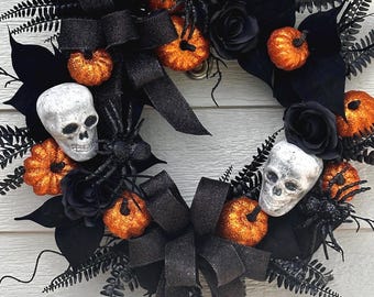May include: A Halloween wreath featuring orange glitter pumpkins, black spiders, and skulls. The wreath is decorated with black roses, fern leaves, and large black bows. The wreath is a festive decoration for the Halloween season.