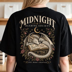 May include: A black t-shirt featuring a graphic on the back with the text MIDNIGHT READING SOCIETY and ONE MORE CHAPTER. The design includes a white dragon curled around an open book, surrounded by floral vines, a crescent moon, and stars.