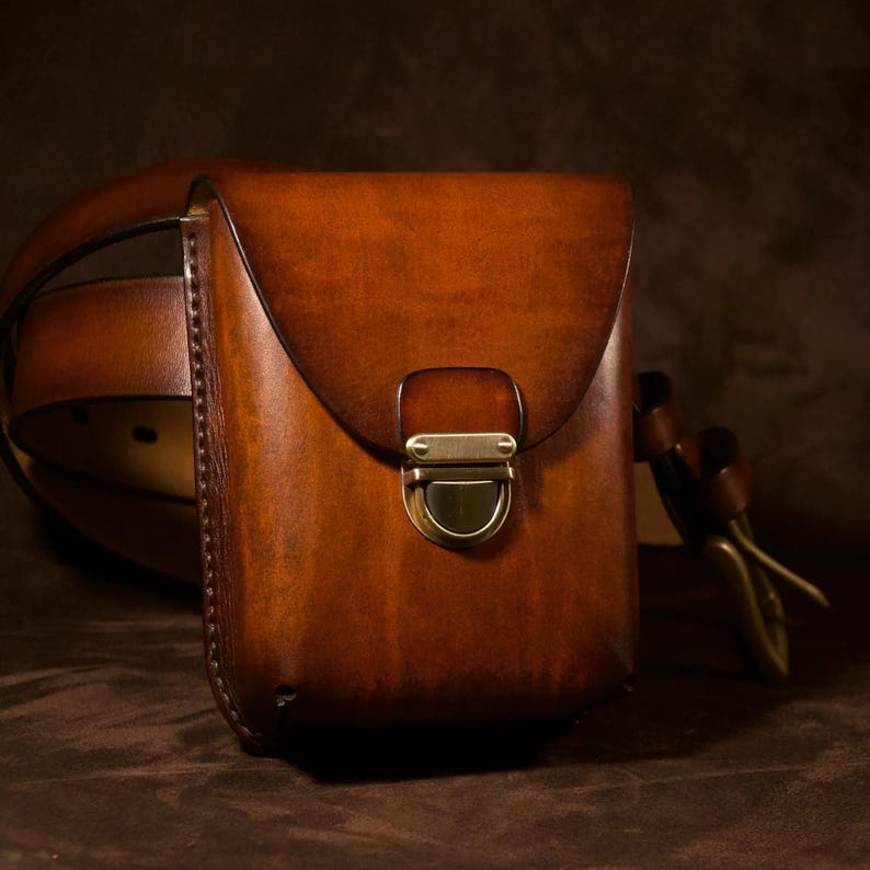 May include: A brown leather belt bag with a flap closure and a brass-colored clasp. The bag is attached to a brown leather belt. The leather has a rich, warm tone, and the bag appears to be handcrafted.