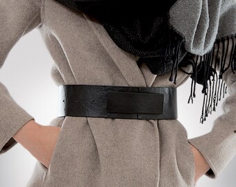 May include: A beige wool coat with a black leather belt. The coat has a shawl collar and is worn open. The belt is wide and has a buckle.