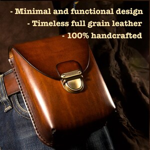 May include: A brown leather belt pouch with a brass clasp, attached to a brown leather belt and blue jeans. The pouch has a curved top and is labeled "- Minimal and functional design - Timeless full grain leather - 100% handcrafted."
