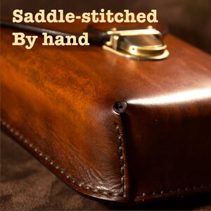 May include: Close-up of a brown leather case with a brass clasp. The text "Saddle-stitched By hand" is visible. The leather has a rich, glossy finish and is hand-stitched with visible thread.