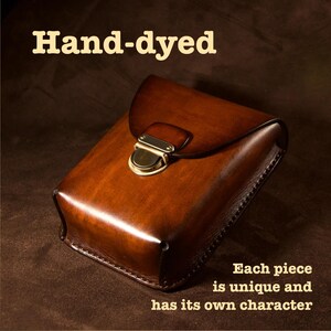 May include: A hand-dyed, brown leather case with a flap closure and brass hardware. The text "Hand-dyed" is at the top, and "Each piece is unique and has its own character" is at the bottom. The leather has a rich, warm tone.