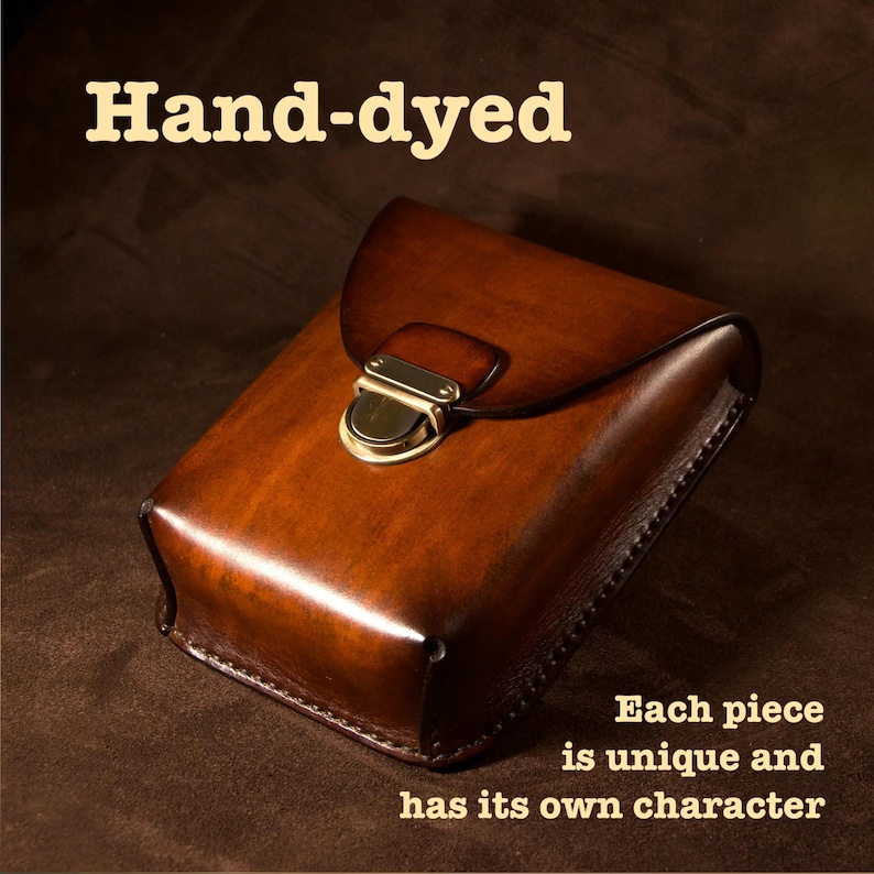 May include: A hand-dyed, brown leather case with a flap closure and brass hardware. The text "Hand-dyed" is at the top, and "Each piece is unique and has its own character" is at the bottom. The leather has a rich, warm tone.