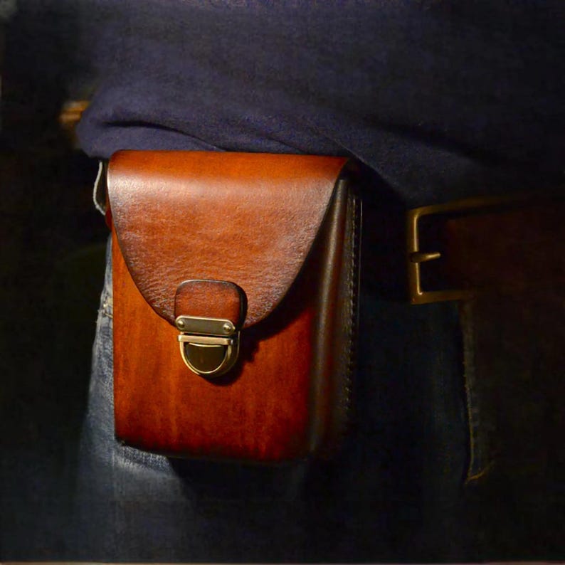 May include: A brown leather pouch with a flap closure and a brass-colored clasp. The pouch is attached to a belt, suggesting it's designed to be worn. The leather has a rich, warm tone, and the design is simple and functional.
