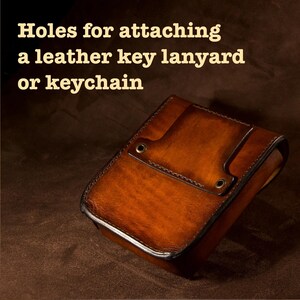 May include: A brown leather case with a flap and two holes for attaching a key lanyard or keychain. The case has a rich, textured appearance and is likely handmade. The text "Holes for attaching a leather key lanyard or keychain" is visible.