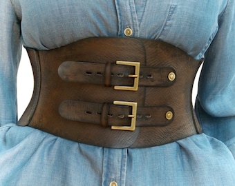 May include: A brown leather corset with two double buckles and gold-toned hardware. The corset is worn over a light blue denim shirt.
