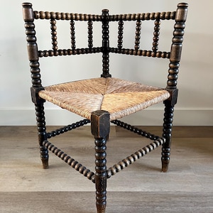 Può includere: Sedia angolare marrone scuro con seduta in paglia intrecciata. La sedia presenta fusi torniti sullo schienale e sulle gambe e una seduta a forma di diamante. La sedia è in legno e ha un aspetto rustico e antico.