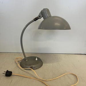 May include: A vintage gray desk lamp with a curved arm and a rounded shade. The lamp has a circular base with a switch and a power cord with a black plug. The lamp is made of metal and has a classic design.