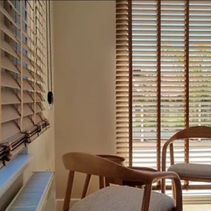 May include: Two wooden chairs with light brown cushions sit in front of a window with wooden blinds. The blinds are open, revealing a view of green trees and a blue sky.