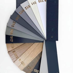 Può includere: Un ventaglio di campioni di colore con varie tonalità di grigio, bianco e legno. Ogni campione è etichettato con un numero in lettere dorate. Il ventaglio è tenuto insieme da un supporto blu scuro.
