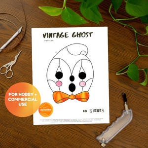 Może przedstawiać: Biała kartka z ilustracją wzoru "Vintage Ghost", przedstawiająca wzór ducha z czarnymi oczami, różowymi policzkami i pomarańczową muszką. Obraz zawiera narzędzia rzemieślnicze i okrągłą naklejkę z napisem "For Hobby + Commercial Use".