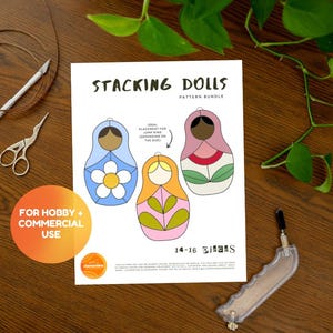 May include: A white paper with the text "STACKING DOLLS PATTERN BUNDLE" and illustrations of three colorful nesting dolls. The dolls are blue, orange, and pink, with floral and leaf designs. The image includes craft tools and a plant.