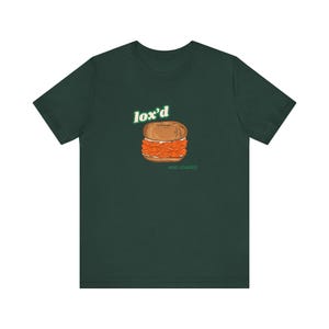 May include: A dark green t-shirt with a graphic of a bagel with lox and cream cheese. The text "lox'd and loaded" is printed below the bagel.