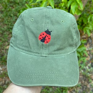 Puede incluir: Una gorra de béisbol verde con una mariquita roja y negra bordada en la parte delantera.