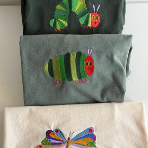 Puede incluir: Tres camisas dobladas con dise&ntilde;os bordados. Dos camisas son de color verde oscuro y gris, con una oruga verde, roja y amarilla. La camisa inferior es de color crema con un dise&ntilde;o de mariposa colorida.