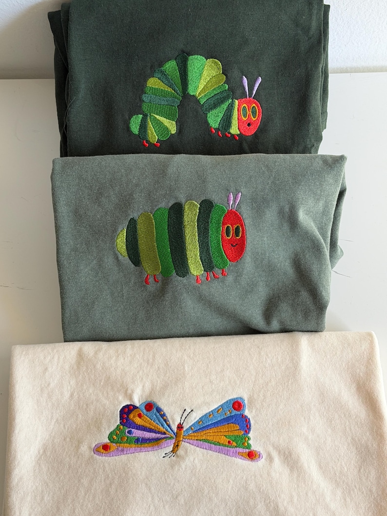 Puede incluir: Tres camisas dobladas con dise&ntilde;os bordados. Dos camisas son de color verde oscuro y gris, con una oruga verde, roja y amarilla. La camisa inferior es de color crema con un dise&ntilde;o de mariposa colorida.