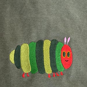 Puede incluir: Dise&ntilde;o de oruga bordada en verde, amarillo y verde oscuro sobre una tela verde oliva apagada. La oruga tiene una cabeza roja con antenas rosas y peque&ntilde;os pies rojos. El dise&ntilde;o est&aacute; centrado.