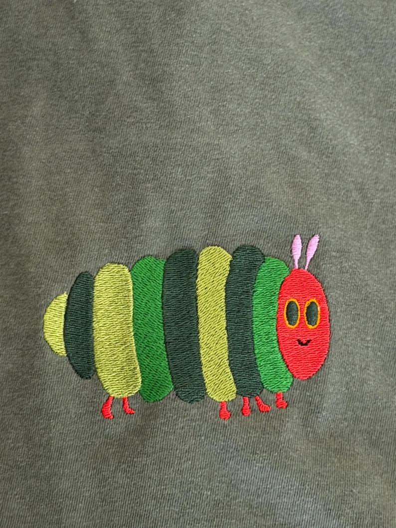 Puede incluir: Dise&ntilde;o de oruga bordada en verde, amarillo y verde oscuro sobre una tela verde oliva apagada. La oruga tiene una cabeza roja con antenas rosas y peque&ntilde;os pies rojos. El dise&ntilde;o est&aacute; centrado.