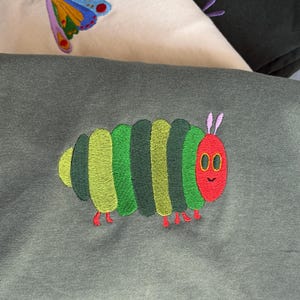 Puede incluir: Primer plano de tres sudaderas con dise&ntilde;os bordados. Una presenta una mariposa colorida, otra una hoja verde y la tercera una oruga verde, roja y amarilla. Las sudaderas son en tonos verdes y crema.