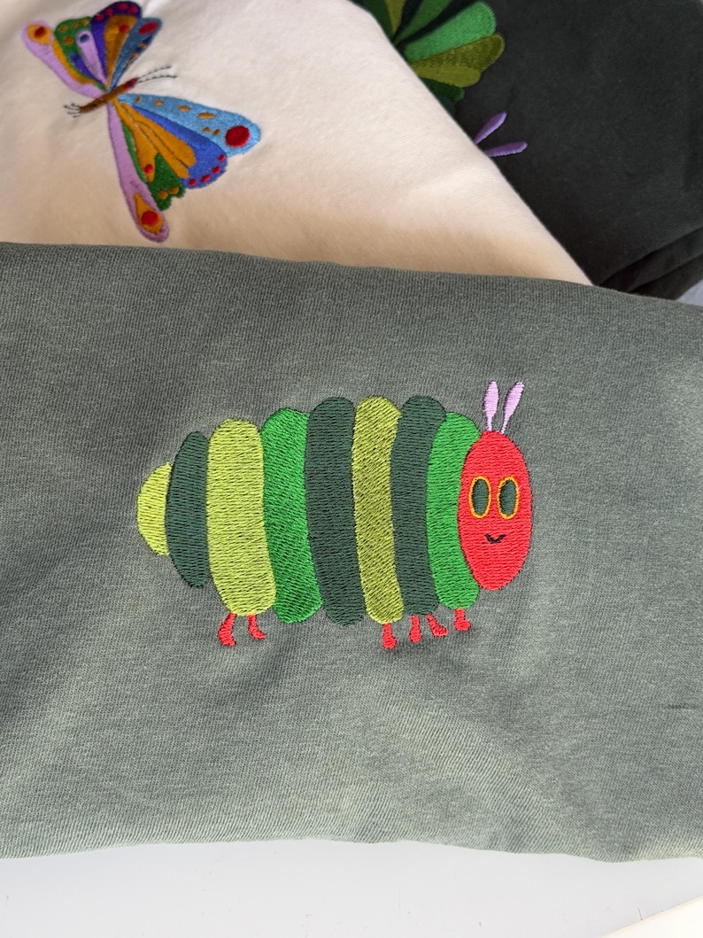 Puede incluir: Primer plano de tres sudaderas con dise&ntilde;os bordados. Una presenta una mariposa colorida, otra una hoja verde y la tercera una oruga verde, roja y amarilla. Las sudaderas son en tonos verdes y crema.