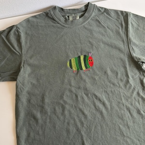 Puede incluir: Una camiseta verde salvia con un dise&ntilde;o de oruga bordada. La oruga tiene una cabeza roja, segmentos corporales verdes y amarillos y patas rojas. La camiseta tiene cuello redondo y mangas cortas.