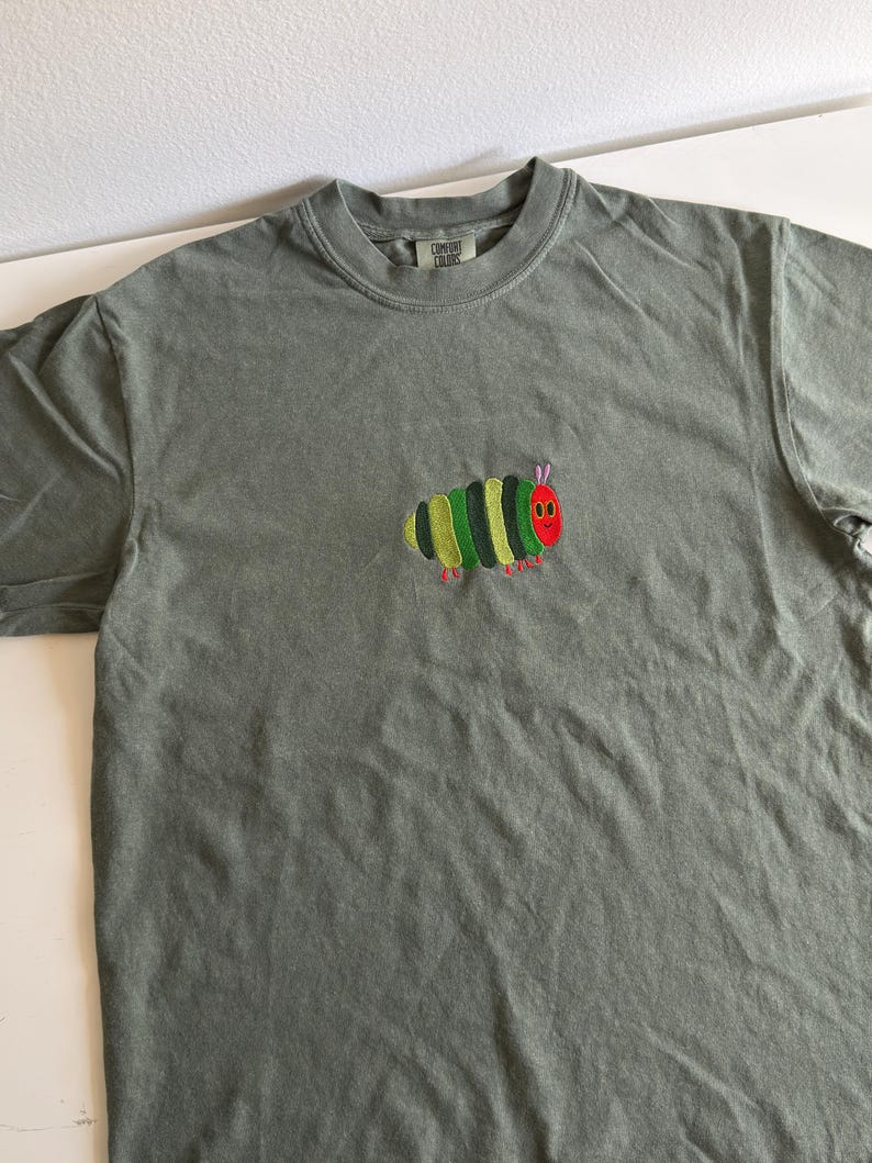 Puede incluir: Una camiseta verde salvia con un dise&ntilde;o de oruga bordada. La oruga tiene una cabeza roja, segmentos corporales verdes y amarillos y patas rojas. La camiseta tiene cuello redondo y mangas cortas.