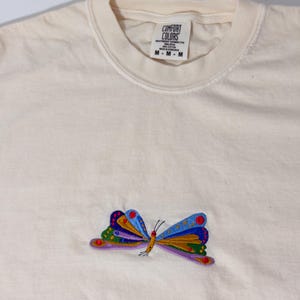May include: Cream-colored t-shirt with a colorful embroidered butterfly design. The butterfly features red, blue, green, and purple details. The shirt has a crew neck and a tag that reads "COMFORT COLORS".