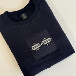 Puede incluir: Una sudadera azul marino oscuro con un parche cuadrado con un diseño de onda blanca estilizada. La sudadera está doblada, mostrando el cuello redondo y el parche bordado. La tela parece suave y cómoda.