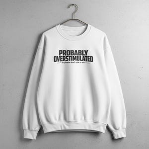 May include: White crewneck sweatshirt with the text "PROBABLY OVERSTIMULATED - so please don't talk to me" in black. The sweatshirt is hanging on a metal hanger against a gray wall.
