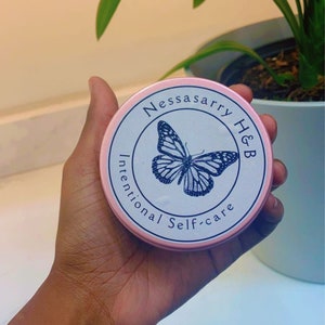 May include: A pink circular container with a white label featuring a butterfly illustration and the text "Nessasarry H&B Intentional Self-care". The container is held in a hand, with a plant in the background.
