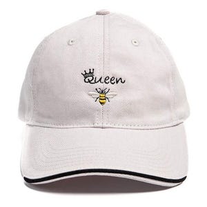 May include: A light gray baseball cap with a black brim. The cap has an embroidered design of a bee with a crown and the word "Queen".