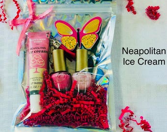 Può includere: Una bustina olografica contenente un lucidalabbra Bath & Body Works Neapolitan Ice Cream, due flaconi di smalto con tappi dorati e un portachiavi a forma di farfalla rosa e gialla. La bustina è legata con un nastro rosa e riempita di carta rossa.