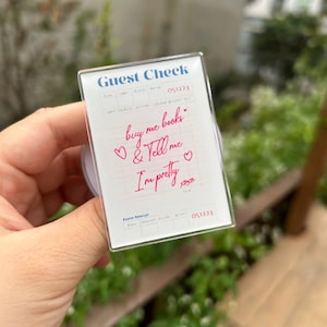 May include: A clear acrylic Guest Check with the words "buy me books & Tell me I'm pretty" in pink script. The top of the check reads "Guest Check" in blue text. The check is held by a hand.