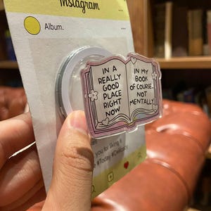 May include: A clear phone grip with a pink book design. The book reads "IN A REALLY GOOD PLACE RIGHT NOW" and "IN MY BOOK OF COURSE... NOT MENTALLY". The grip is attached to a yellow Instagram-themed backing.