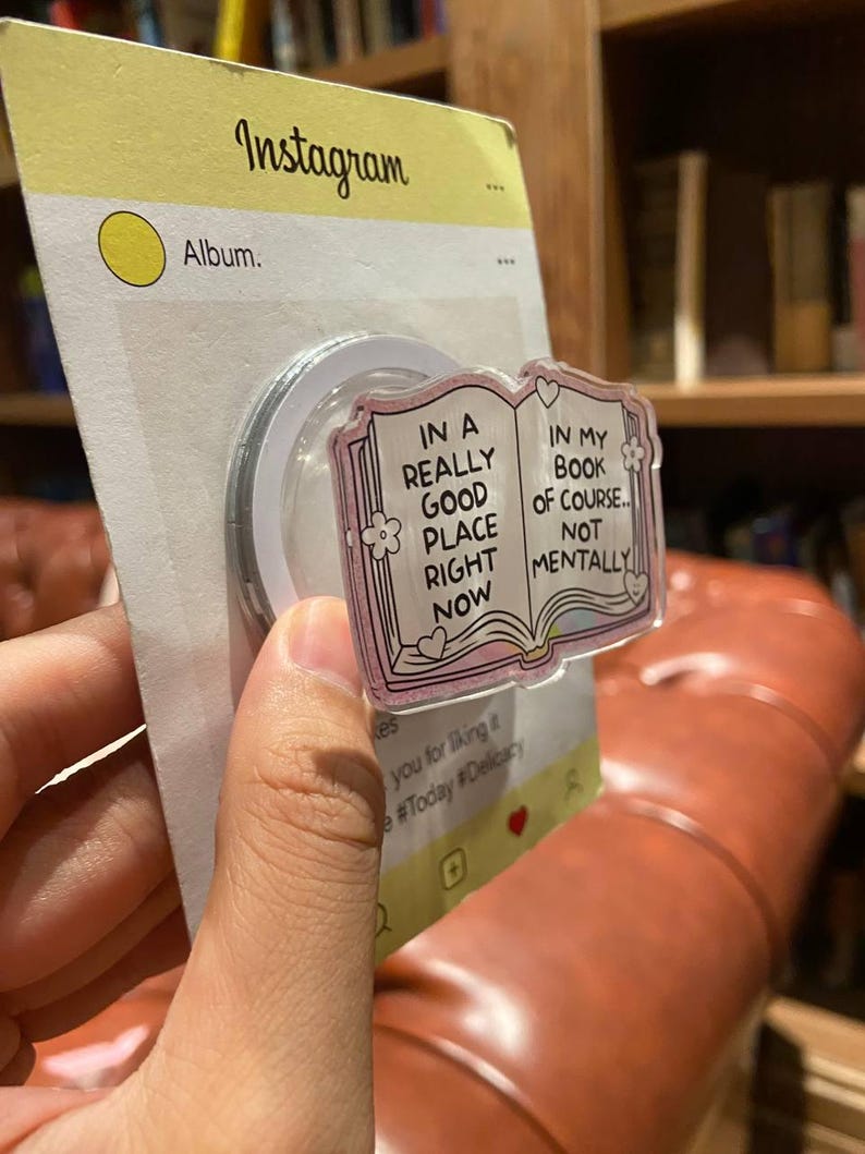 May include: A clear phone grip with a pink book design. The book reads "IN A REALLY GOOD PLACE RIGHT NOW" and "IN MY BOOK OF COURSE... NOT MENTALLY". The grip is attached to a yellow Instagram-themed backing.