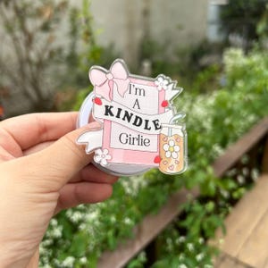 May include: A clear acrylic sticker with the text "I'm A KINDLE Girlie" in black and pink. The sticker features a pink bow, flowers, and a drink with a flower design.