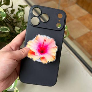 May include: A matte black smartphone case featuring a circular collapsible grip attached to the back. The grip displays a vibrant, detailed illustration of a hibiscus flower with bright pink, orange, and yellow petals and a prominent central stamen.