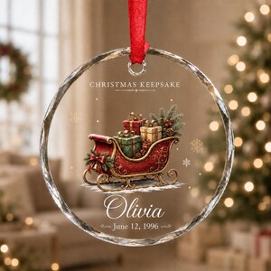 May include: A circular glass Christmas ornament with a red ribbon hanger. The design features a red sleigh filled with wrapped gifts, pine branches, and pinecones. Text reads: CHRISTMAS KEEPSAKE, Olivia, and June 12, 1996, with gold snowflakes.