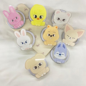 May include: A collection of eight circular phone grips featuring cartoon animal characters including a pink rabbit, yellow chick, white cat, white rabbit, tan hamster, beige fennec fox, light blue wolf, and a tan puppy, all arranged on a cream display card.