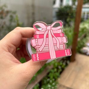 May include: A pink and red stack of books with a pink bow on top. The books are in varying shades of pink and red. The bow has long ribbons that drape down the front of the books. The image is a clear, glossy sticker.