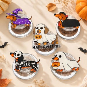 May include: Five circular phone grips featuring cartoon dachshunds in Halloween costumes: a purple witch, a black vampire, a ghost with a pumpkin bucket, a skeleton, and a ghost with a pumpkin bucket above the text HALLOWEENIE.