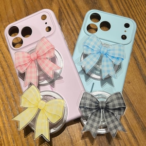 May include: Two smartphone cases in light pink and light blue, each featuring two circular phone grips. The grips are decorated with acrylic bow charms in a gingham pattern, available in pink, blue, yellow, and grey color variations.